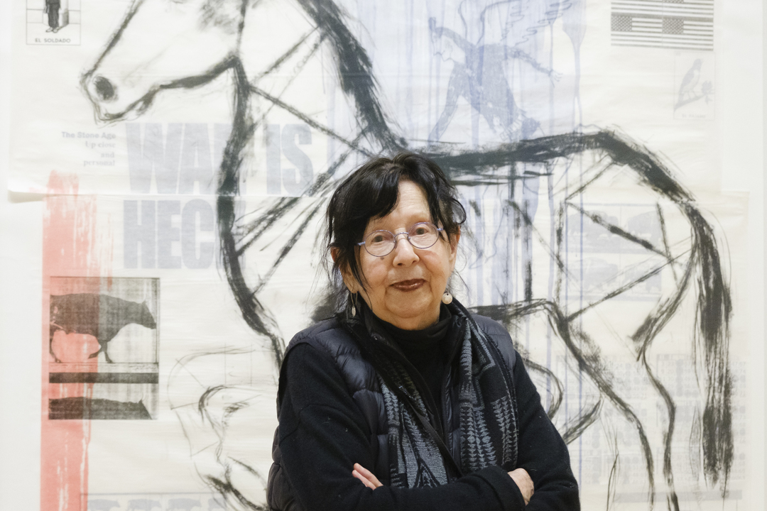 An older woman with glasses and dark hair stands confidently, arms crossed, in a black jacket and scarf. Behind her, a large abstract artwork featuring a horse is displayed on the wall—a tribute to artist and activist Jaune Quick-to-See Smith.