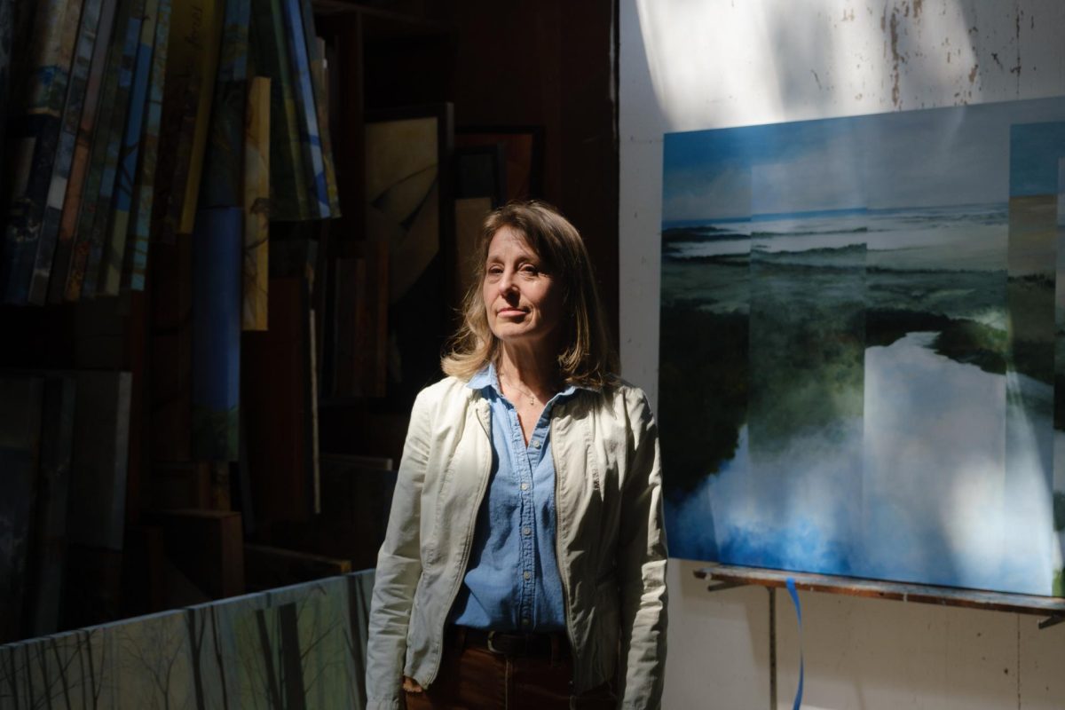 A woman in a white jacket and denim shirt stands in a dimly lit room with paintings around her. Sunlight streams in, highlighting her face and some artwork.