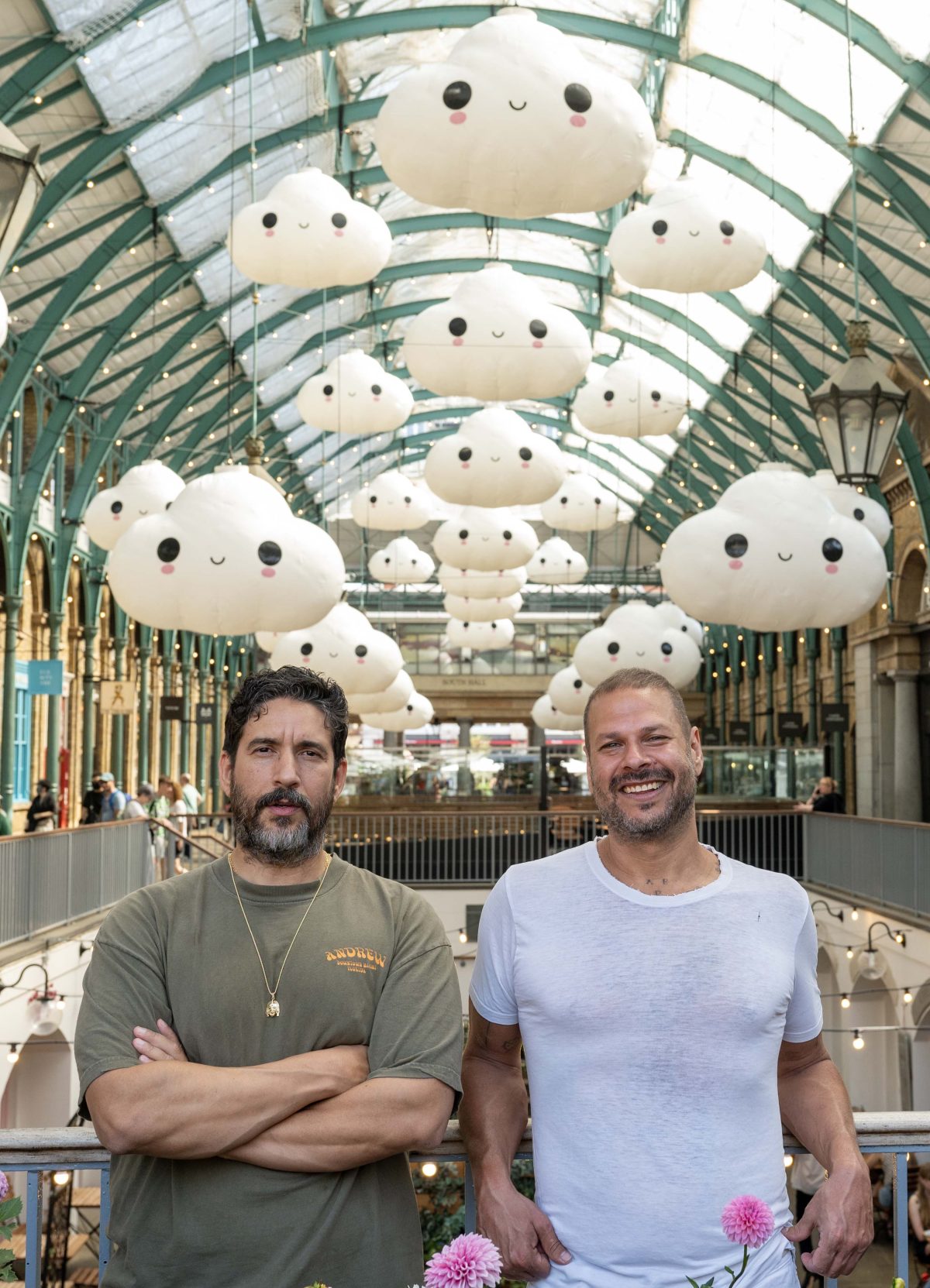 Two people stand in an indoor market, under adorable inflatable cloud decorations by FriendsWithYou. The market features a glass ceiling and iron supports. Both individuals are smiling, dressed casually, with pink flowers adding a splash of color below.